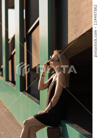 Young woman in a black dress, talking on a cell phone, walking on a city street in the summertime 114497862