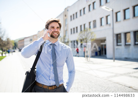 Handsome businessman phone calling on smartphone, standing in city street. Working remotely, waiting for business meeting. Manager smiling, outdoor in urban setting 114498633
