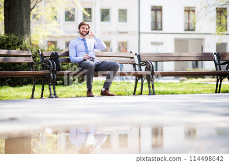 Handsome businessman phone calling on smartphone, sitting on city bench, writing notes. Working remotely, waiting for business meeting. Manager smiling, outdoor in urban setting Handsome businessman phone calling on smartphone, sitting on city bench, writing notes. Working remotely, waiting for business meeting. Manager smiling, outdoor in urban setting 114498642