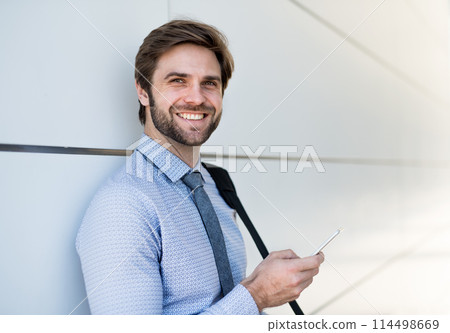 Handsome businessman scrolling on smartphone standing in front office building going to work. Manager smiling, outdoor in urban setting Handsome businessman scrolling on smartphone standing in front office building going to work. Manager smiling, outdoor in urban setting 114498669