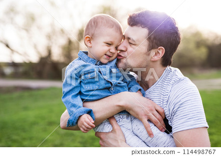 Father holding little toddler boy kissing him, making him laugh, having fun during warm spring day. Father's day concept. 114498767