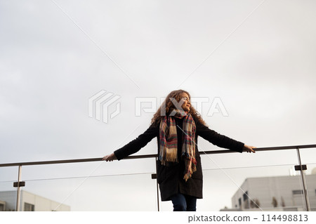 Beautiful woman with curly hair standing on terrace, enjoying cold autumn morning, sunny day. Speaking positive affirmations. 114498813