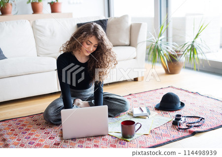 Beautiful woman planning summer vacation abroad, going on trip alone. Sitting on floor, working on itinerary on laptop. 114498939