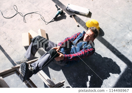 Male worker lying on the floor after fall from ladder, holding his knee. Work injury, accident in workplace. Male worker lying on the floor after fall from ladder, holding his knee. Work injury, accident in workplace. 114498967