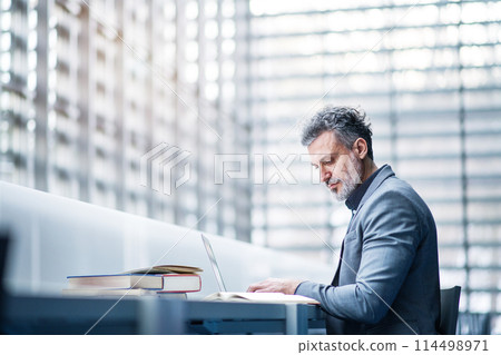 Handsome businessman working on laptop, searching for informations in book. Traditional information source in modern office space. 114498971
