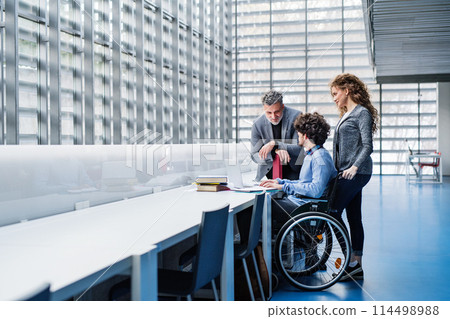 Handsome university professor helping student in wheelchair, showing him book for his thesis in library. Handsome university professor helping student in wheelchair, showing him book for his thesis in library. 114498988