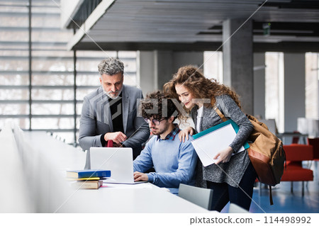 Handsome university professor helping to student final project, showing him research paper, book for thesis in library. Handsome university professor helping to student final project, showing him research paper, book for thesis in library. 114498992