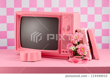A pink television placed in center of photo with some lifestyle accessories and a blank pedestal in flower shaped placed in front, against light pink checkered background. Frontal shot photo A pink television placed in center of photo with some lifestyle accessories and a blank pedestal in flower shaped placed in front, against light pink checkered background. Frontal shot photo 114499010