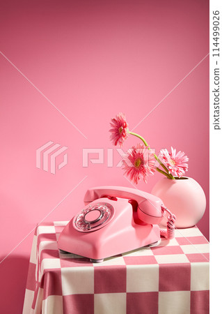 Pink background against a desk phone with pink color and a flower vase of pink gerbera daisy placed on a table has checkered pattern. Frontal shot with space for text 114499026