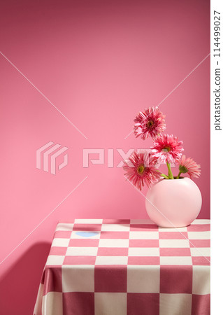 Advertising photo on pink background. A checkered table containing a white vase holding some pink gerbera rosa flower in corner. Photo was from front view with blank space for design Advertising photo on pink background. A checkered table containing a white vase holding some pink gerbera rosa flower in corner. Photo was from front view with blank space for design 114499027