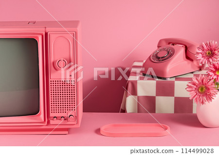A pink table containing an empty tray in the center with television on the left side and a white vase of pink gerbera rosa decorated on the other side, a desk phone on checkered pink table behind 114499028