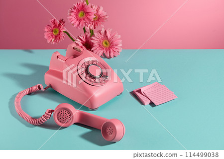 A desk phone with receiver placed on a flat blue surface, a few branches of pink gerbera flowers decorated on the back and a stack of sticky notes of the same color placed next to it. Copy space 114499038