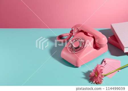 Pink color desk phone, some books, sticky notes and a gerbera daisy branch displayed on the right side of blue table over pink background. Blank space on the left for showing product or adding text 114499039