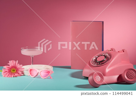 Life style pink item on blue table over pink background, a desk phone, pink pedestal, a glasses, cocktail glass and daisy gerbera. Space for adding text, front view 114499041