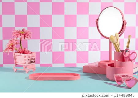Stationery items in pink color neatly arranged in the corner of blue table, an empty tray placed in center over checkered pink background. Frontal shot photo with empty space for presentation 114499043