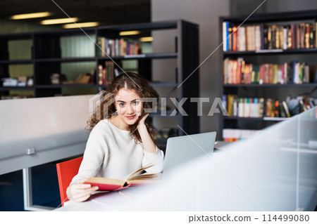 Young female student in library, focusing on final project, presentation. University student preparing for final exam. 114499080
