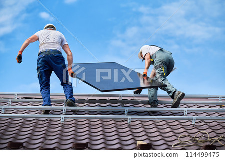 Workers building solar panel system on roof of house. Two men installers in helmets carrying photovoltaic solar module outdoors. Alternative, green and renewable energy generation concept. 114499475