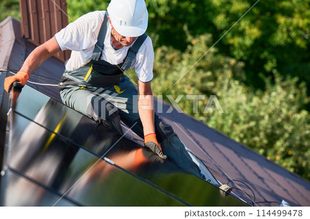 Worker building solar panel system on rooftop of house. Man engineer in helmet installing photovoltaic solar module outdoors. Alternative, green and renewable energy generation concept. Worker building solar panel system on rooftop of house. Man engineer in helmet installing photovoltaic solar module outdoors. Alternative, green and renewable energy generation concept. 114499478