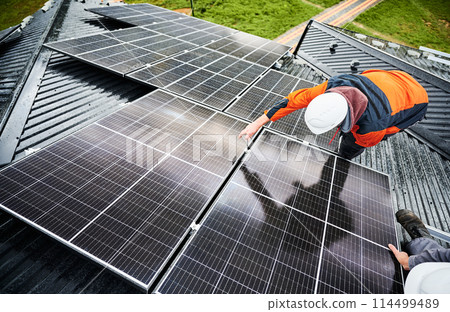 Man builder mounting photovoltaic solar panels on roof of house. Engineer in helmet installing solar module system with help of hex key. Concept of alternative, renewable energy. 114499489