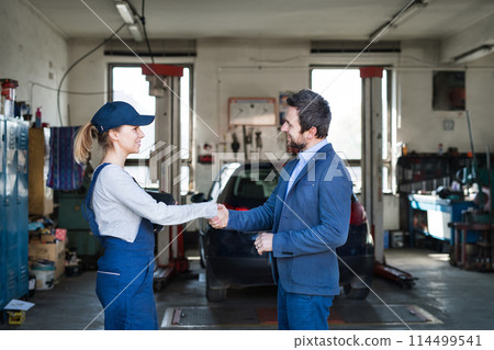 Female auto mechanic talking with customer, shaking hands. Beautiful woman working in a garage, wearing blue coveralls. Female auto mechanic talking with customer, shaking hands. Beautiful woman working in a garage, wearing blue coveralls. 114499541