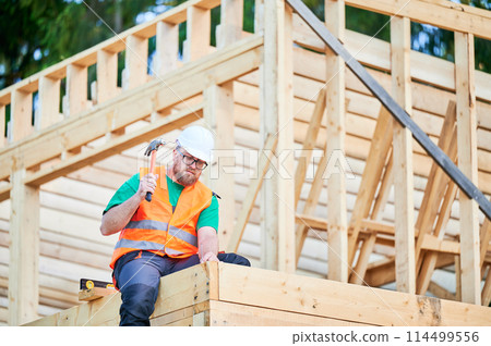 Carpenter constructing two-story wooden frame house. Bearded man in glasses hammering nails into structure while wearing protective helmet, construction vest. Concept of modern ecological construction 114499556