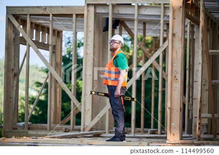 Carpenter building wooden frame two-story house. Bearded man in glasses holding a level, wearing protective helmet, overalls and orange vest. Concept of modern ecological construction. 114499560