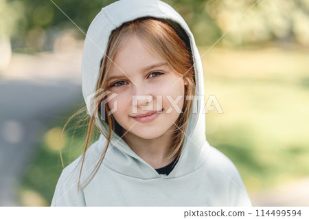 Portrait of happy little girl in hoodie smiling at camera while walking in park on blurred nature background 114499594