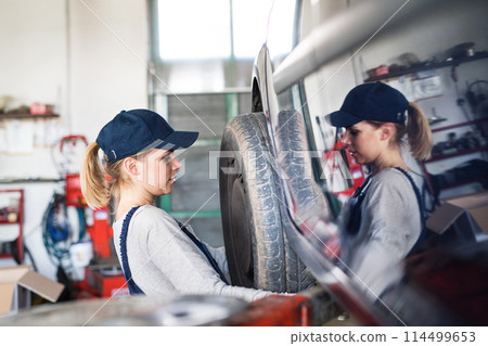 Female auto mechanic changing tieres in auto service. Beautiful woman holding tire in a garage, wearing blue coveralls. 114499653