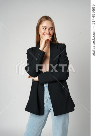 Young Woman in Casual Blazer and Jeans Smiling Against a Grey Background Young Woman in Casual Blazer and Jeans Smiling Against a Grey Background 114499699