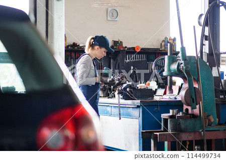 Female auto mechanic repairing, maintaining car. Beautiful woman working in a garage, wearing blue coveralls. 114499734
