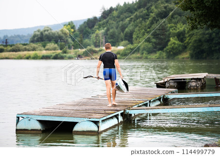 Young canoeist carry canoe and paddle, going into water, walking on wooden dock. Concept of canoeing as dynamic and adventurous sport Young canoeist carry canoe and paddle, going into water, walking on wooden dock. Concept of canoeing as dynamic and adventurous sport 114499779