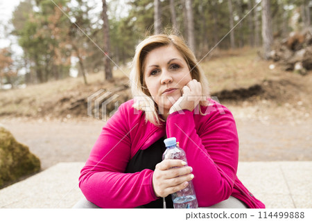 Overweight woman drinking water after run in nature. Exercising outdoors for people with obesity. Overweight woman drinking water after run in nature. Exercising outdoors for people with obesity. 114499888