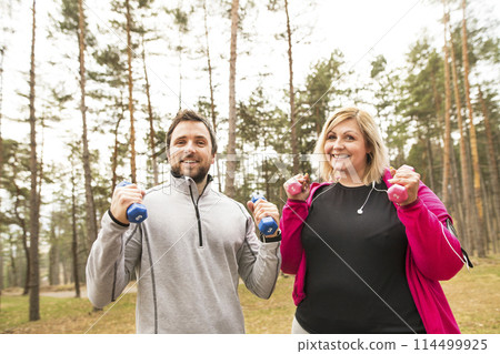 An overweight woman exercising outdoors with friend, fitness coach. Holding dumbbells. 114499925