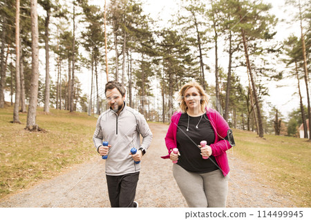 An overweight woman running in nature with friend. Exercising outdoors for people with obesity, support from friend, fitness coach. An overweight woman running in nature with friend. Exercising outdoors for people with obesity, support from friend, fitness coach. 114499945
