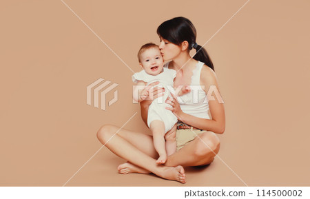 Happy young mother holding and kissing baby on studio background 114500002