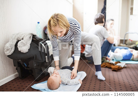 Mother changing diaper after group exercise class. Moms staying active, working out with baby in gym, bonding. 114500005