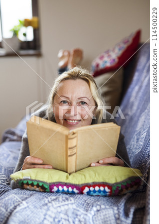 Beautiful mature woman reading book, feeling cozy and happy. Weekend activity for older woman, relaxing at home. 114500229