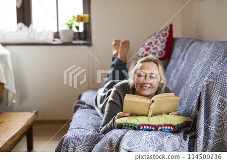 Beautiful mature woman reading book, feeling cozy and happy. Weekend activity for older woman, relaxing at home. 114500236