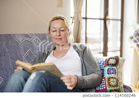 Beautiful mature woman reading book, feeling cozy and happy. Weekend activity for older woman, relaxing at home. 114500246
