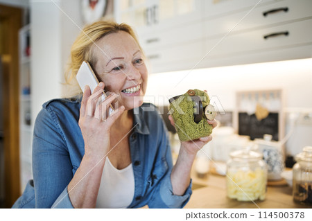 Beautiful mature woman at home, phone calling and drinking hot herbal tea. Older woman living alone, enjoying peaceful weekend day 114500378