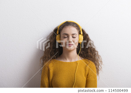 Portrait of a gorgeous teenage girl with curly hair, listening music via headphones. Studio shot, white background with copy space 114500499