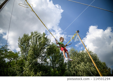 The boy is jumping on a bungee trampoline. A child with insurance and stretchable rubber bands hangs against the sky. The concept of happy childhood and games in the amusement park The boy is jumping on a bungee trampoline. A child with insurance and stretchable rubber bands hangs against the sky. The concept of happy childhood and games in the amusement park 114501294