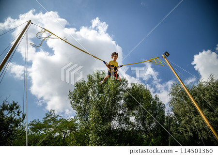 The boy is jumping on a bungee trampoline. A child with insurance and stretchable rubber bands hangs against the sky. The concept of happy childhood and games in the amusement park The boy is jumping on a bungee trampoline. A child with insurance and stretchable rubber bands hangs against the sky. The concept of happy childhood and games in the amusement park 114501360