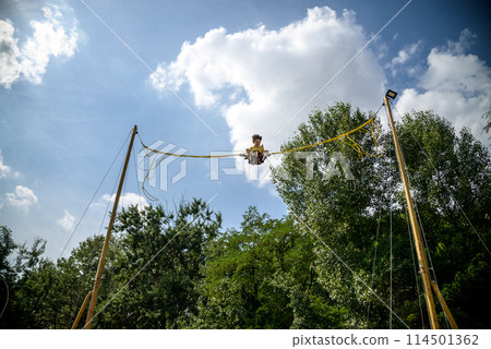 The boy is jumping on a bungee trampoline. A child with insurance and stretchable rubber bands hangs against the sky. The concept of happy childhood and games in the amusement park 114501362