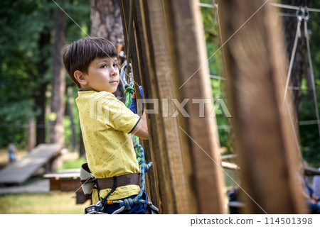 Strong excited young boy playing outdoors in rope park. Caucasian child dressed in casual clothes and sneakers at warm sunny day. Active leisure time with children concept Strong excited young boy playing outdoors in rope park. Caucasian child dressed in casual clothes and sneakers at warm sunny day. Active leisure time with children concept 114501398