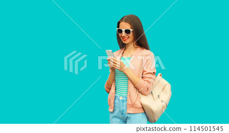 Portrait of smiling young woman with smartphone wearing backpack and sunglasses on blue background 114501545