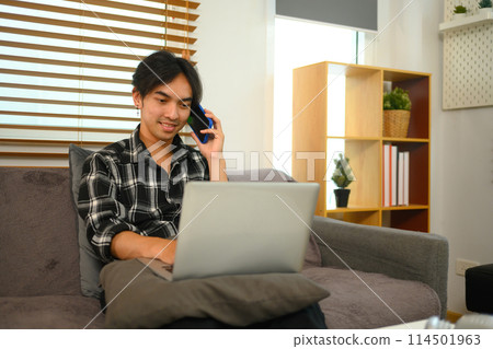 Smiling young man having phone conversation and working with laptop at home 114501963