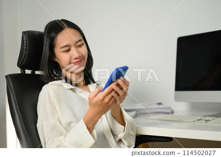 Smiling female office worker using mobile phone at her workplace 114502027