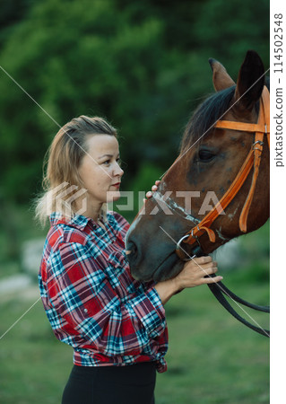 Happy blonde with horse in forest. Woman and a horse walking through the field during the day. Dressed in a plaid shirt and black leggings. Happy blonde with horse in forest. Woman and a horse walking through the field during the day. Dressed in a plaid shirt and black leggings. 114502548