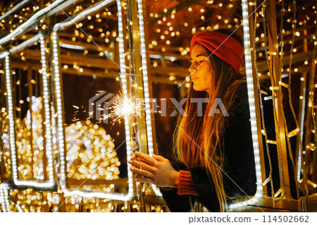 Woman holding sparkler night while celebrating Christmas outside. Dressed in a fur coat and a red headband. Blurred christmas decorations in the background. Selective focus 114502662